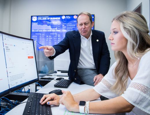 A student sitting at a computer with a male faculty member teaching her