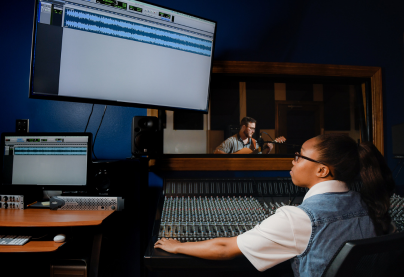 female student working at the mixing board while a male student plays guitar in the recording studio