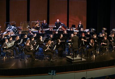 A large university concert band performs on stage, with musicians playing woodwinds, brass, and percussion under the direction of a conductor.