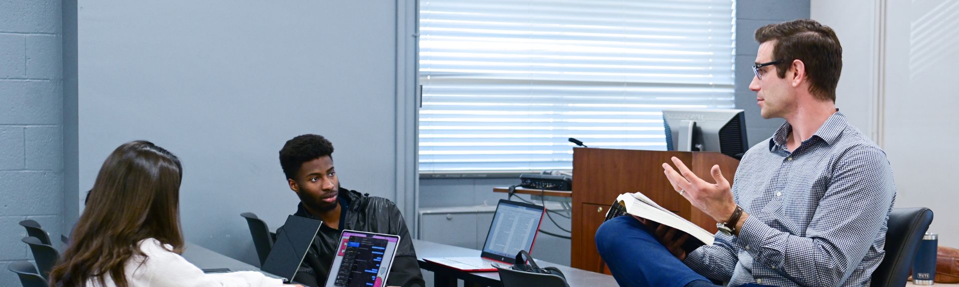 A professor sits in a chair holding an open book and gestures while speaking to a small group of students seated around tables with laptops in a classroom.